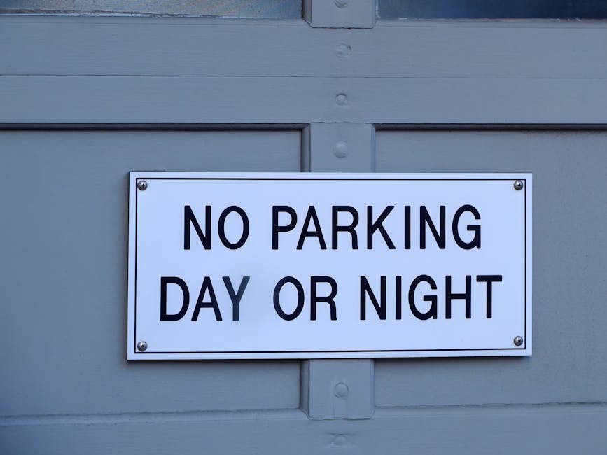 A round no parking traffic sign with a red border, blue interior, and a red diagonal line across it is mounted on a grey metal pole set against a textured brick wall composed of irregular cream, beige, and light brown stones separated by mortar, with a horizontal band of reddish-brown bricks running across the lower part of the wall. The sign casts a shadow onto the wall, indicating sunlight from the right side of the image. The scene appears to be outdoors, potentially in a residential or commercial area, highlighting parking restrictions that could impact home relocation or furniture transport processes managed by Removal Companies Hounslow. The detailed brickwork and the sign's visibility suggest considerations for parking regulations during house removals, ensuring compliance with local parking and skip rules as outlined under the Hounslow Council policies.