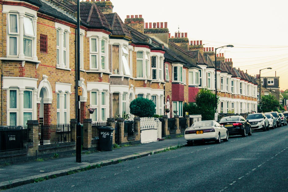A row of traditional terraced houses with brick facades and white bay windows lining a residential street during daylight. The pavement in front features a black metal fence, small front gardens, and some potted plants. Several vehicles, including a white car, a black car, and a silver van, are parked along the street. The scene captures a typical urban environment suitable for home relocation or furniture transport, with streetlights and utility poles visible. This image is relevant to house removals and moving logistics, illustrating the typical environment where professional removals, such as those provided by Removal Companies Hounslow, operate to assist clients with packing, loading, and transport services in the area.