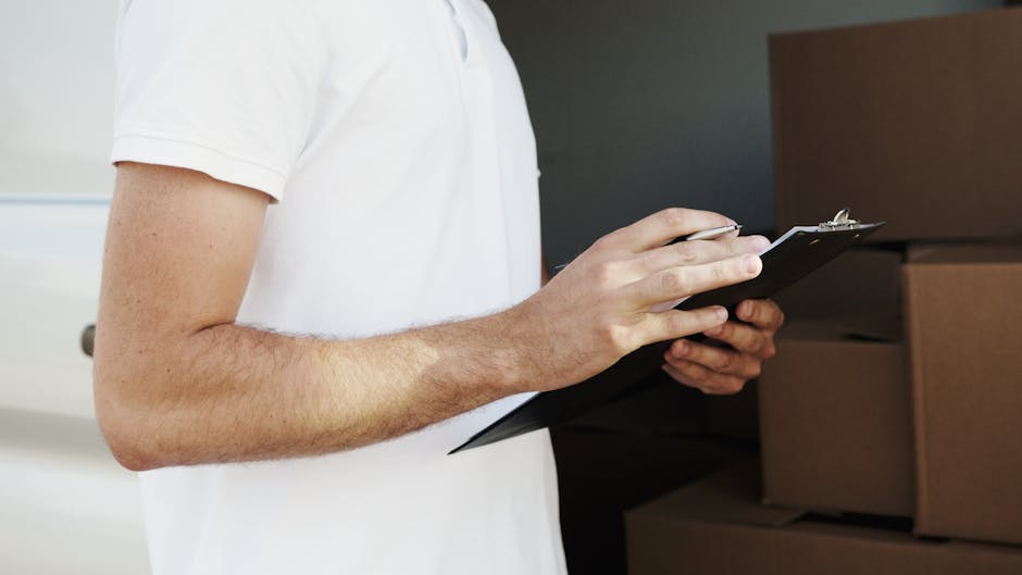 A person wearing a white t-shirt holds a black clipboard with a pen, standing next to several packed cardboard boxes and storage containers stacked inside an indoor space. The boxes appear to be prepared for a house move, with some wrapped in plastic for protection, and are situated near a doorway with natural lighting illuminating the area. The scene depicts the process of organizing or managing a home relocation, aligning with furniture transport and packing and moving activities. The individual is possibly a representative from Removal Companies Hounslow, involved in overseeing the loading process or verifying items for house removals in preparation for transportation to a new property.
