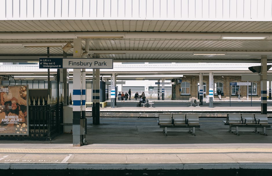 A view of Finsbury Park train station platform showing several passengers seated on metal benches under a large, white, corrugated steel canopy supported by grey metal columns. A sign with the station name, 'Finsbury Park,' is prominently displayed above the platform. The platform surface is made of concrete, with tactile paving near the edge for accessibility. To the left, there is a poster advertising a theatrical production, and behind the platform, multiple train tracks are visible with a few people walking along the platform or waiting. The station environment is well-lit with natural daylight, and the overall scene reflects a typical urban railway station setting, which could be relevant for house removals if the context involves transporting household items via train or planning access for moving vehicles, as managed by Removal Companies Hounslow.