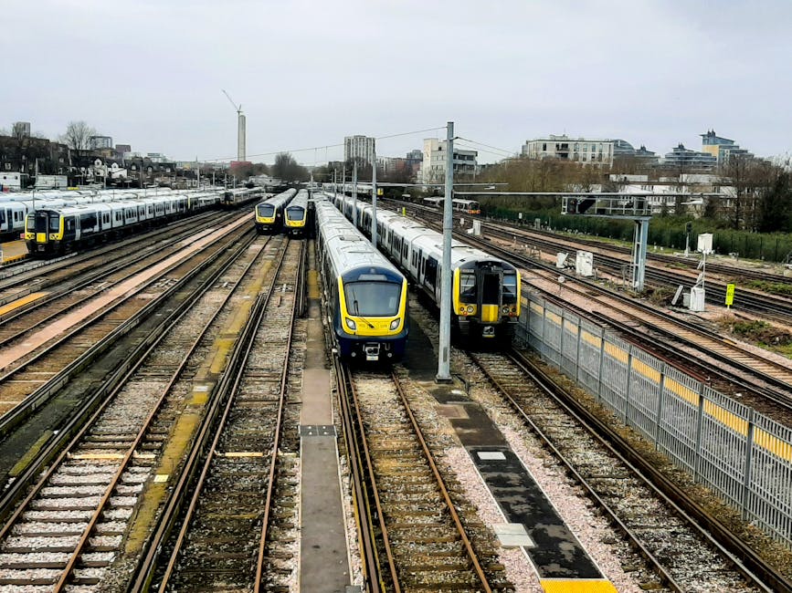 A view of multiple train carriages parked on railway tracks in a train depot or station, with two modern yellow and grey trains positioned side by side in the central area, and additional trains visible in the background on adjacent tracks. The scene includes overhead electrical lines supported by metal poles, a paved walkway running alongside the tracks, and a distant cityscape with buildings and trees under an overcast sky. The image captures a broad perspective of the railway yard, highlighting the extensive network of tracks and the alignment of the trains, which are part of a home relocation or furniture transport service connected to removals activities by Removal Companies Hounslow. The environment suggests an industrial area adapted for logistics, with no visible human activity but emphasizing the scale of train movement and loading infrastructure.