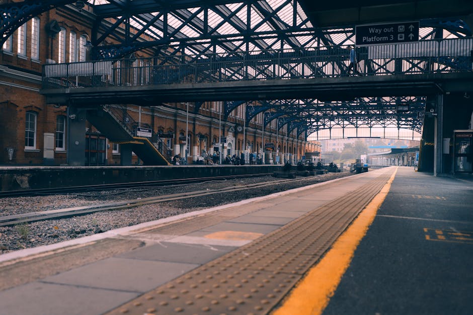 A view of Finsbury Park train station platform showing several passengers seated on metal benches under a large, white, corrugated steel canopy supported by grey metal columns. A sign with the station name, 'Finsbury Park,' is prominently displayed above the platform. The platform surface is made of concrete, with tactile paving near the edge for accessibility. To the left, there is a poster advertising a theatrical production, and behind the platform, multiple train tracks are visible with a few people walking along the platform or waiting. The station environment is well-lit with natural daylight, and the overall scene reflects a typical urban railway station setting, which could be relevant for house removals if the context involves transporting household items via train or planning access for moving vehicles, as managed by Removal Companies Hounslow.