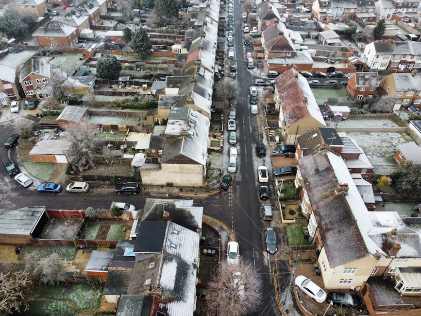 An aerial view of a residential area on Lampton Road showing narrow pavements and access challenges for house removals in Hounslow. The street is lined with closely spaced terraced houses and semi-detached homes, some with small front gardens and driveways filled with parked cars. The road surface appears wet, indicating recent rain, and is obstructed in some areas by vehicles, including vans and cars parked along the curbs. Several houses have visible roof structures with tiles, and backyards enclosed by fences and garden sheds. A removal van from Removal Companies Hounslow is parked on the street near one of the houses, with furniture and boxes possibly inside, while a trolley and packing materials are situated on the pavement, indicating ongoing packing and loading activities. The scene illustrates typical home relocation logistical considerations in tight urban spaces, where access problems on Lampton Road may require careful planning for efficient furniture transport, packing, and moving process coordination.