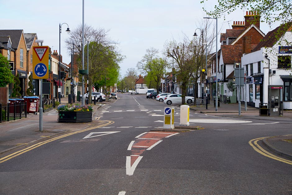 A quiet residential street with a central roundabout and a 'GIVE WAY' traffic sign. The scene includes parked cars along both sides of the street, small shops and buildings with traditional brick facades on the right, and a lamppost with a decorative flower planter on the left. Trees with budding green leaves line the pavements, indicating early spring. The road features painted markings such as a dashed red box junction and directional arrows guiding vehicles around the roundabout. Street lighting and utility poles are visible, with clear, natural daylight illuminating the scene. This image reflects an urban environment suitable for home relocation activities, with available space for moving vans and container loading, and is relevant to the logistics of furniture transport during house removals. The setting is calm and organized, typical of a suburban area where [COMPANY_NAME] might conduct a house move or assist with access arrangements during a removal process, highlighting typical logistical considerations for removals services on roads with access constraints as described in the 'Lampton Road access problems for Hounslow removals' page.