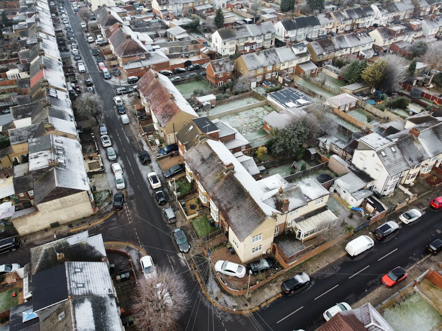 An aerial view of a residential area on Lampton Road showing narrow pavements and access challenges for house removals in Hounslow. The street is lined with closely spaced terraced houses and semi-detached homes, some with small front gardens and driveways filled with parked cars. The road surface appears wet, indicating recent rain, and is obstructed in some areas by vehicles, including vans and cars parked along the curbs. Several houses have visible roof structures with tiles, and backyards enclosed by fences and garden sheds. A removal van from Removal Companies Hounslow is parked on the street near one of the houses, with furniture and boxes possibly inside, while a trolley and packing materials are situated on the pavement, indicating ongoing packing and loading activities. The scene illustrates typical home relocation logistical considerations in tight urban spaces, where access problems on Lampton Road may require careful planning for efficient furniture transport, packing, and moving process coordination.