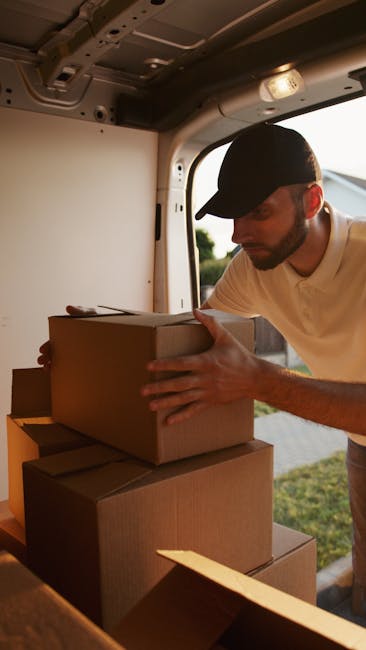 A man wearing a black cap and white t-shirt is inside a vehicle, possibly a van used for home relocation, carefully stacking or adjusting cardboard boxes during the loading process. The boxes are brown and vary in size, with some being placed on top of others inside the vehicle's interior, which features a ceiling with visible fixtures and a window showing an outdoor scene with greenery. The man is partially illuminated by natural light from the open side door or back of the van, highlighting his focused effort in secure packing or furniture transport. Removal Companies Hounslow, a professional removal service, appears to facilitate this logistics and packing operation, supporting home removals or furniture transport needs in the area.