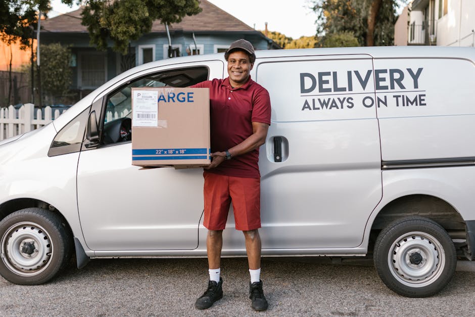A smiling delivery person from Removal Companies Hounslow stands outside a white delivery van during a home relocation process, holding a medium-sized cardboard box labeled with handling and delivery details. The van is parked on a residential street with houses, trees, and a fence visible in the background. The vehicle's side door is closed, and the company branding, including the words 'DELIVERY ALWAYS ON TIME,' is prominently displayed. The delivery person is dressed in a red polo shirt, red shorts, black sneakers, and a grey cap, ready to load or unload items as part of packing and moving services. The scene captures the efficient loading process with the individual preparing to transport household items, highlighting the logistical steps involved in furniture transport and home relocation, managed professionally by [COMPANY_NAME]. The environment is well-lit, indicating daytime, with a focus on the practical aspects of removals and moving logistics.