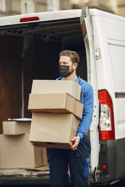 A man wearing a black cap and white t-shirt is inside a vehicle, possibly a van used for home relocation, carefully stacking or adjusting cardboard boxes during the loading process. The boxes are brown and vary in size, with some being placed on top of others inside the vehicle's interior, which features a ceiling with visible fixtures and a window showing an outdoor scene with greenery. The man is partially illuminated by natural light from the open side door or back of the van, highlighting his focused effort in secure packing or furniture transport. Removal Companies Hounslow, a professional removal service, appears to facilitate this logistics and packing operation, supporting home removals or furniture transport needs in the area.
