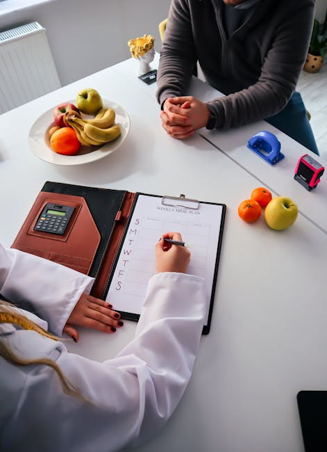 The image shows two individuals seated at a white table inside a room, engaged in planning a home relocation. One person, dressed in a white coat, is writing on a weekly planner with days of the week listed vertically, while the other, wearing a dark long-sleeved top, has their hands clasped on the table. On the table, there is a bowl of assorted fresh fruits including bananas, apples, and oranges, along with two small tape measures—one with a pink case and another with a blue case—suggesting packing or measurement activities. Visible nearby are cardboard boxes with packing materials, possibly for furniture or fragile items, indicating the packing process involved in house removals. The background includes a radiator and minimal decor, emphasizing the focus on planning and logistics for an upcoming move. This scene is associated with services provided by Removal Companies Hounslow, specializing in domestic removals and furniture transport.