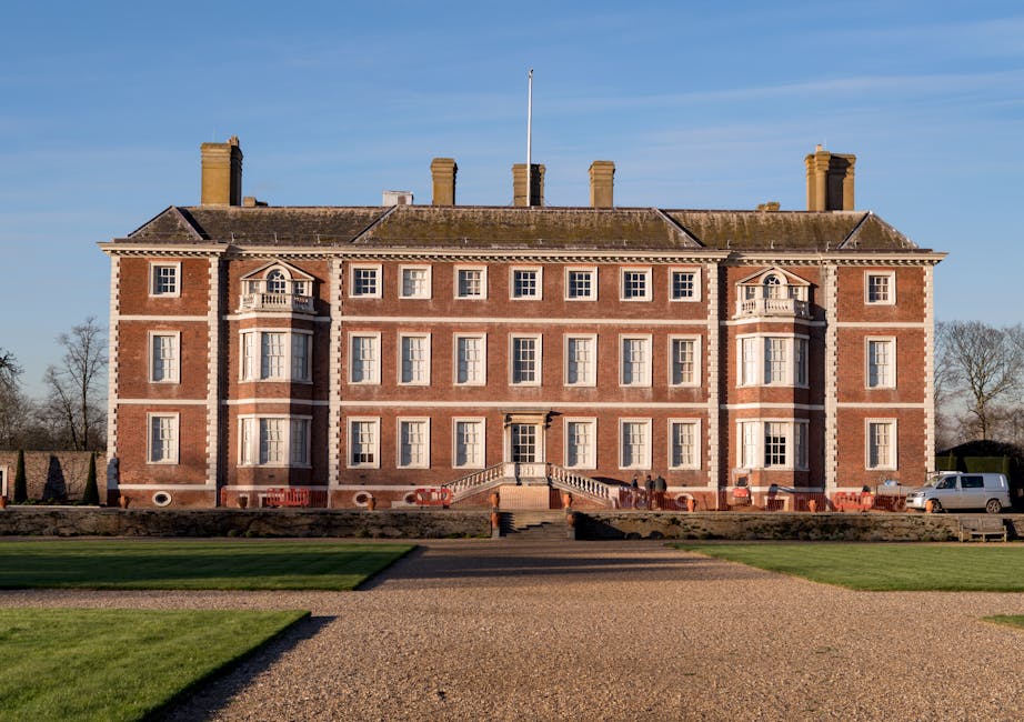 A large historic brick building with four stories, featuring symmetrical windows with white frames, decorative stonework, and small gabled protrusions on the upper corners. In front of the building, a wide stone staircase with metal handrails leads to a central doorway, flanked by evenly spaced landscaping and low brick wall boundaries. The foreground includes a gravel path and well-maintained grassy areas, with a clear blue sky overhead. To the right side of the building, a white van is parked near the pavement, and several orange and white barriers are positioned along the edge of the property indicating ongoing or completed house removals. The setting suggests a location suitable for home relocation services and furniture transport, with the building's spacious exterior providing ample room for loading and unloading of packed boxes, furniture, and appliances as part of a professional removal process, supported by the expertise of Removal Companies Hounslow.
