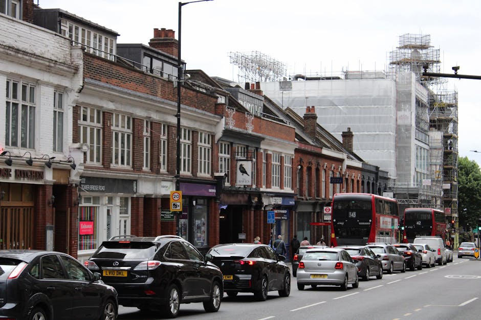 The image depicts a busy street scene on Hounslow High Street during daytime, with a row of traditional brick-fronted shops and office buildings lining the pavement. In the foreground, a line of parked cars, including sedans and hatchbacks, extends along the curb. Several pedestrians are walking on the sidewalk, and a group of people is seen near the shop entrances. Two red double-decker buses are visible, one approaching from the right and the other further down the street, indicating active public transportation. Behind the shops, a building is under construction, covered with scaffolding and white protective sheeting, suggesting ongoing renovation or development work. The street is busy with vehicles and pedestrians, and the overall scene captures a typical urban area involved in moving or home relocation activities, as managed by companies like Removal Companies Hounslow, with active loading and unloading zones possibly involved nearby.