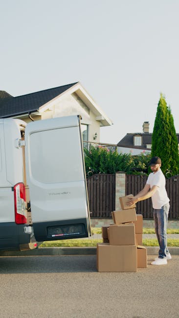 A residential street scene featuring a row of parked vehicles on both sides of the narrow tarmac road, including various vans and cars, some of which are covered with protective materials such as blan