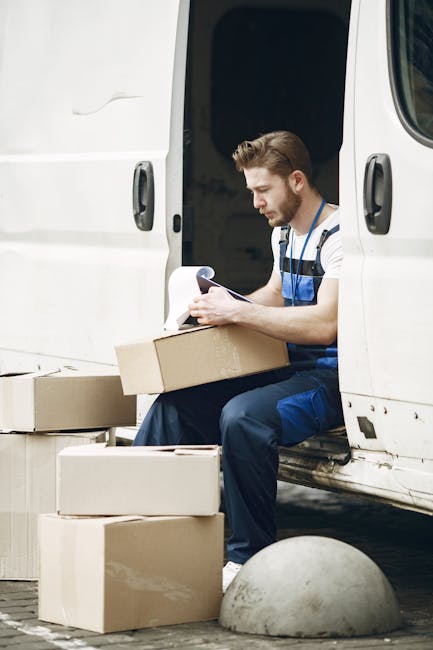 A young male removal worker with short, styled brown hair and a beard, sitting on the edge of an open white delivery van during a home relocation process. He is wearing a blue and white striped shirt with dark blue overalls, and is focused on reading a document or checklist. Surrounding him are several cardboard boxes of various sizes, some stacked on the ground and one in his hands, likely packed for moving. The boxes are made of brown corrugated cardboard, and some are taped shut, consistent with packing materials used in professional removals. The van's interior is visible, showing a dark space suitable for loading furniture and boxes. The setting suggests an outdoor loading area, possibly at a residential property, with the worker preparing for furniture transport and unloading tasks typical of a same-day removals service in Hounslow, as promoted by Removal Companies Hounslow. The scene emphasizes the packing, organizing, and logistics involved in house removals and furniture transport.