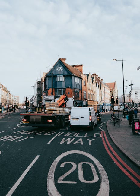 A street scene in Hounslow featuring a delivery or removal truck with a flatbed carrying several large cardboard boxes, wooden pallets, and packing materials, parked in the middle of the road near a pedestrian crosswalk on a clear day. The truck is positioned close to the curb on the left side, adjacent to a white van that appears to be involved in a home relocation or furniture transport process. The surrounding area includes multiple multi-story buildings with brick and painted facades, some with storefronts at street level, and various street lamps and bicycles parked along the sidewalk. The road has marking lines, including a 20-mph speed limit, and the scene suggests an active urban environment suitable for house moves or relocation services, with the company 'Removal Companies Hounslow' possibly conducting or preparing for a loading operation within the city. Bright natural lighting highlights the scene, emphasizing the logistical aspects of loading and unloading in a busy town center. This image visually supports services related to professional removals and furniture transport in Hounslow.
