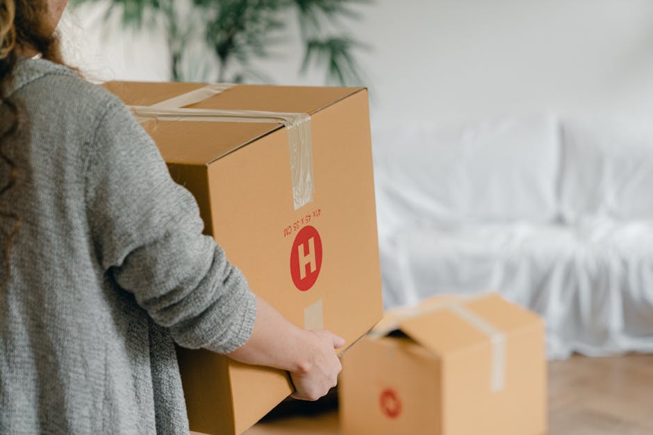 A person wearing a grey sweater is seen holding a large cardboard moving box with one hand, while using a box cutter with the other hand to carefully open the top flaps. The box is marked with red printed symbols and labels, including a prominent red 'H' and weight specifications. In the background, additional unsealed boxes are visible on the floor, suggesting an active packing or packing process during home relocation. The setting appears to be an indoor space with natural lighting, and a white covered sofa is partially visible in the background. The scene illustrates furniture transport and packing activities typical of house removals performed by Removal Companies Hounslow, demonstrating the careful handling and organisation involved in professional removals alongside the loading process involved in moves within or out of the TW3 postcode area.