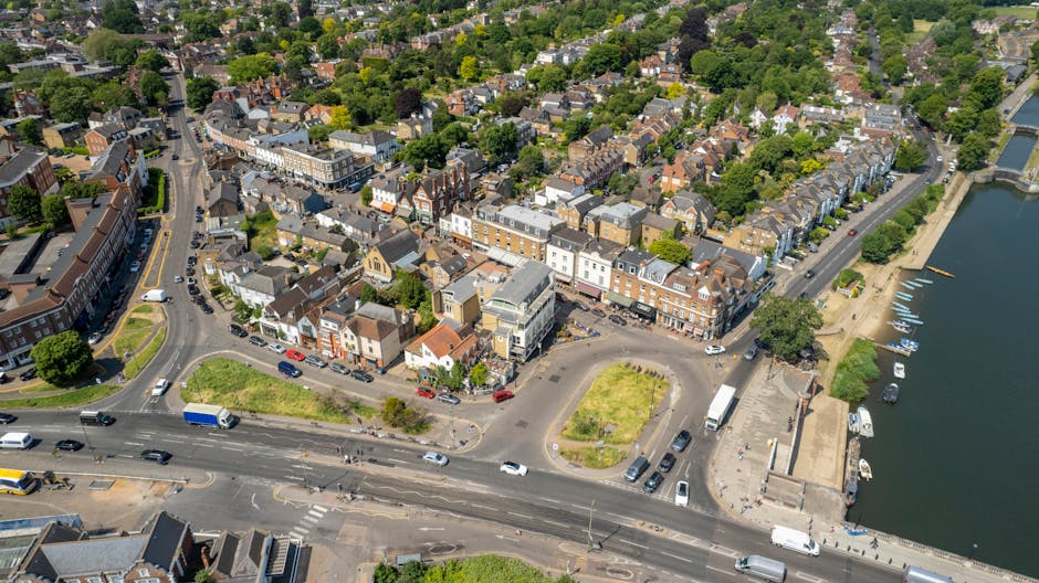 An aerial view of a riverside town showing a mix of commercial and residential buildings, with a busy road running parallel to the water. The scene includes parked cars along the street, a small parking area with trucks and vans likely involved in a home relocation or removals process, and a designated loading zone with furniture and boxes being prepared for transport. Some items, such as cardboard boxes, wrapping materials, and pieces of furniture, are visible in the area near the vehicles, indicating a moving or packing activity. The waterway on the right features boats moored along the quay. Trees and green spaces are scattered throughout the densely built neighbourhood, which includes traditional terraced houses and low-rise commercial premises. The natural lighting suggests daytime, and the environment reflects a typical urban setting suitable for professional removals services, as offered by Removal Companies Hounslow, supporting domestic moving logistics in the TW3 postcode area.