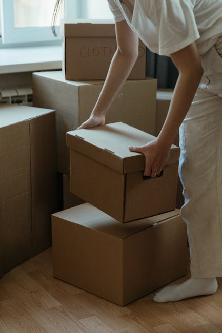 A person wearing a grey sweater is seen holding a large cardboard moving box with one hand, while using a box cutter with the other hand to carefully open the top flaps. The box is marked with red printed symbols and labels, including a prominent red 'H' and weight specifications. In the background, additional unsealed boxes are visible on the floor, suggesting an active packing or packing process during home relocation. The setting appears to be an indoor space with natural lighting, and a white covered sofa is partially visible in the background. The scene illustrates furniture transport and packing activities typical of house removals performed by Removal Companies Hounslow, demonstrating the careful handling and organisation involved in professional removals alongside the loading process involved in moves within or out of the TW3 postcode area.