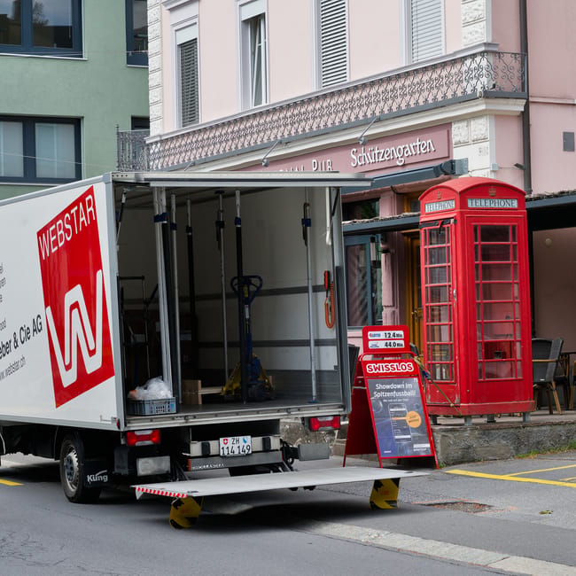 A white moving truck with the logo of Webstair and tracking information on the back is parked on a city street in front of a building, with its cargo area open and empty, showing lifting straps, a hand truck, and packing materials inside. The truck is positioned on a street with a landing area that has small levelers for ease of loading and unloading furniture. Adjacent to the truck, there is a classic red telephone box and a sidewalk with a building featuring a balcony and decorative architectural elements. The scene captures the process of home relocation or furniture transport, with the truck ready for loading or unloading large items such as boxes, appliances, or furniture pieces, supported by professional removals services like those provided by Removal Companies Hounslow.