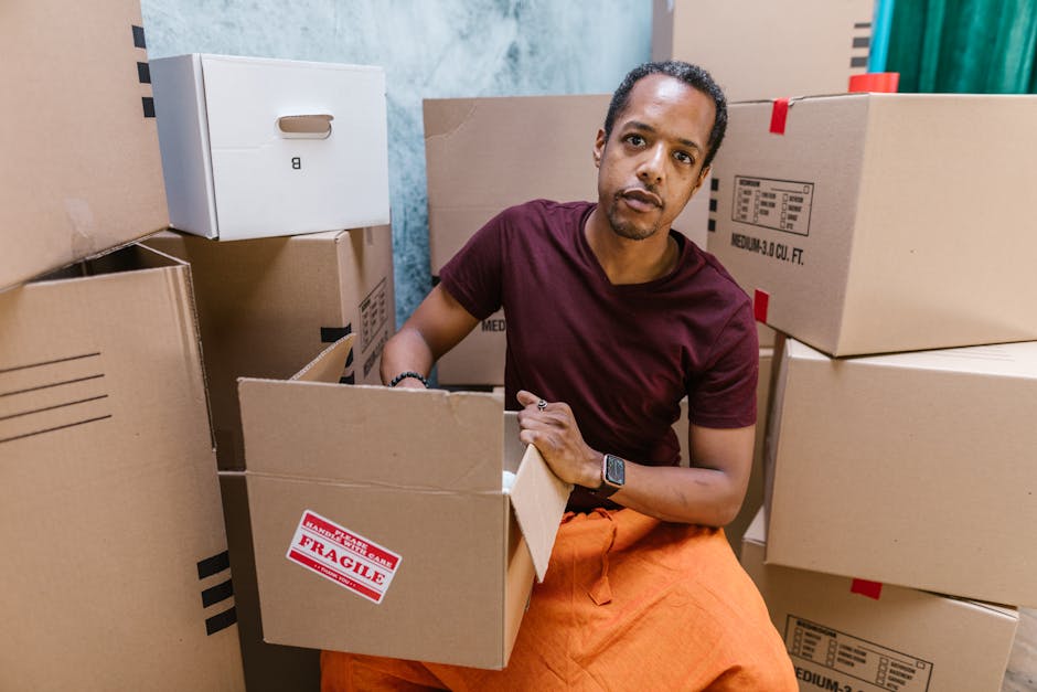 A man wearing a black cap, white polo shirt, and jeans is kneeling on an outdoor pavement during daylight, surrounded by open cardboard boxes filled with packing materials. He is writing on a piece of paper placed on top of one of the boxes, preparing for a home relocation. Behind him, additional boxes are stacked, some sealed with packing tape. The scene shows part of a moving vehicle with an open door to the right, indicating the loading process of furniture or household items. The background includes a blurred road and greenery, suggesting a residential or suburban setting. Removal Companies Hounslow is involved in furniture transport and packing and moving services, as evidenced by the organized loading activity depicted.